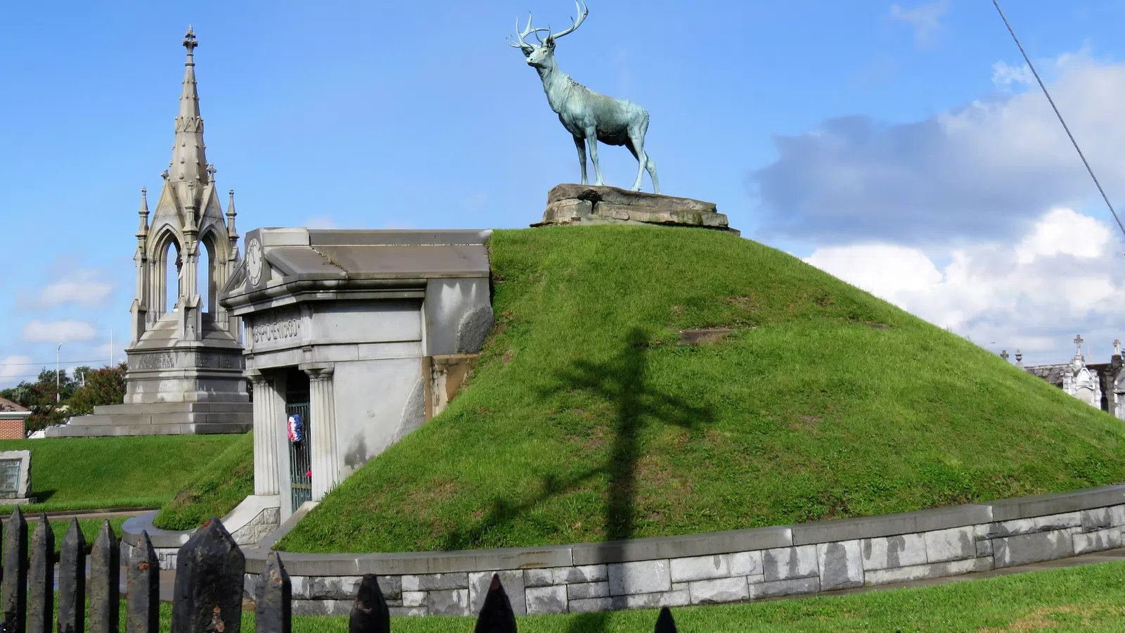 Bronze stag statue atop green grassy mound at historic cemetery under blue sky with scattered clouds.