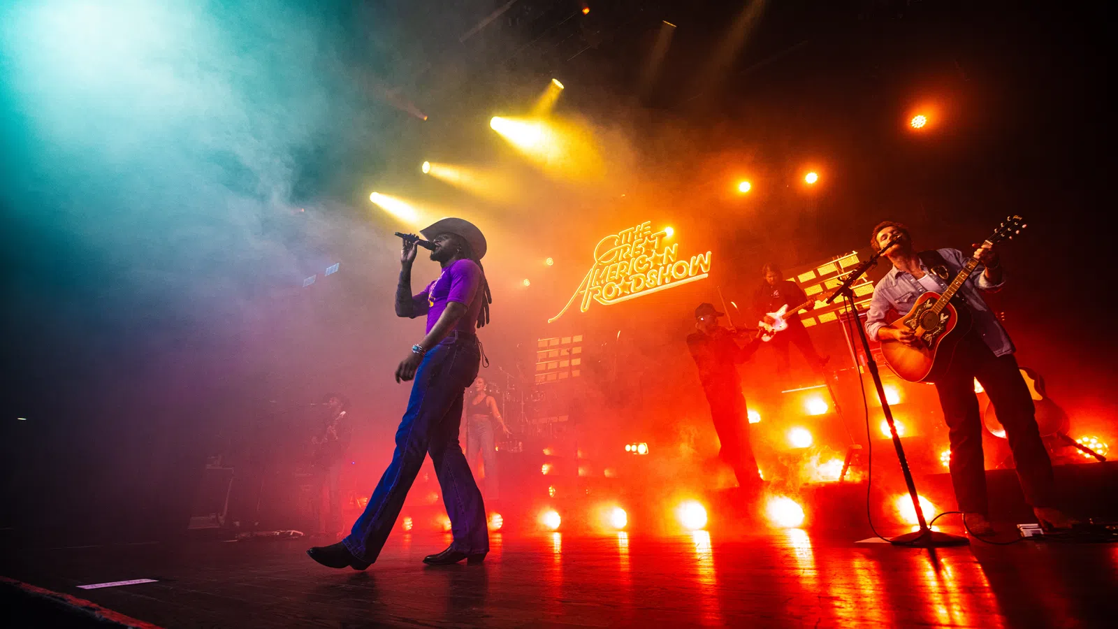 Live concert with a singer in a cowboy hat, guitarist, and vibrant colorful stage lighting and fog effects