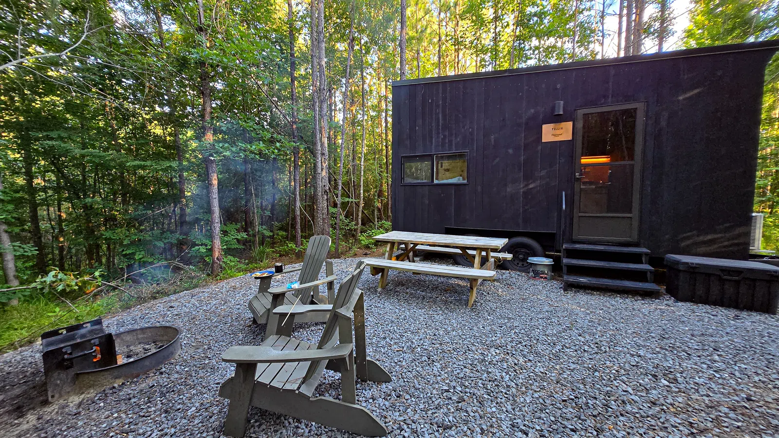 Tiny black cabin in forest clearing with picnic table, Adirondack chairs, and a fire pit on gravel ground.
