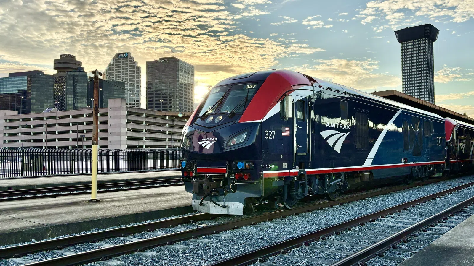 Amtrak train parked on tracks at a station with city skyline and vibrant cloudy sky at sunset.