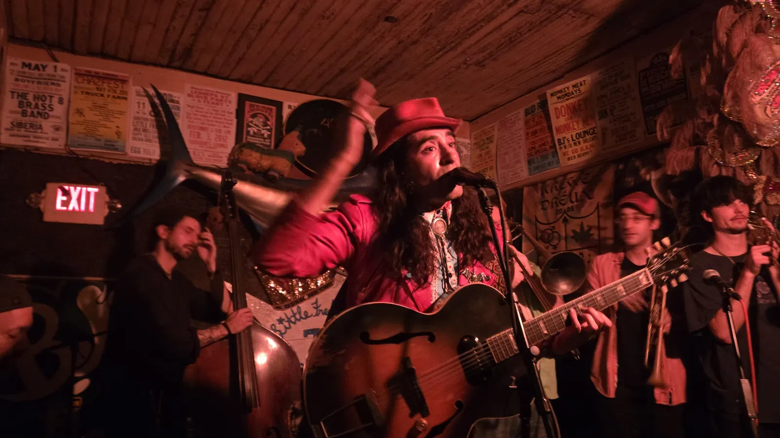 Live band performing in a cozy, dimly lit venue with musician singing and playing guitar surrounded by brass instruments.