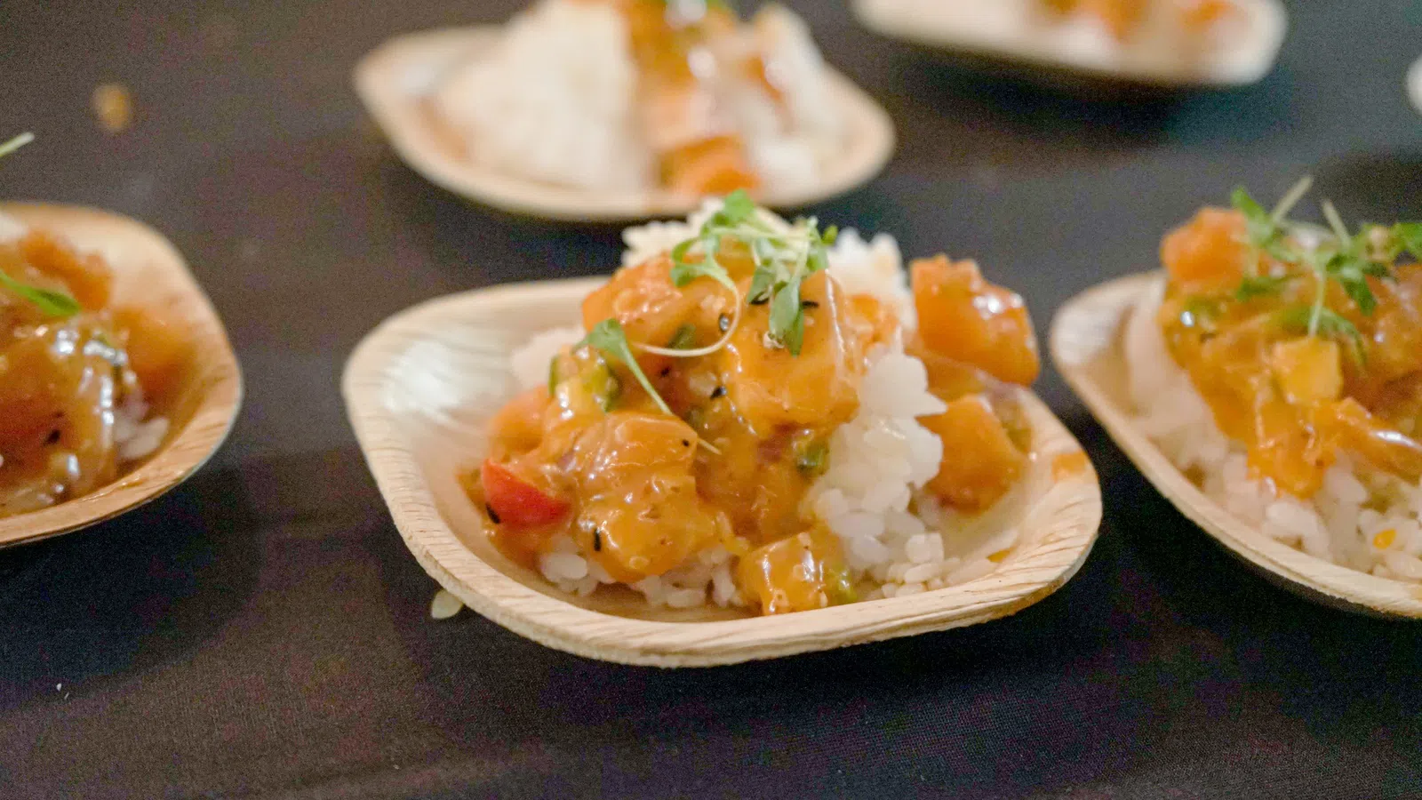 Close-up of seafood poke bowls served on small biodegradable plates with rice and garnished herbs on black table.