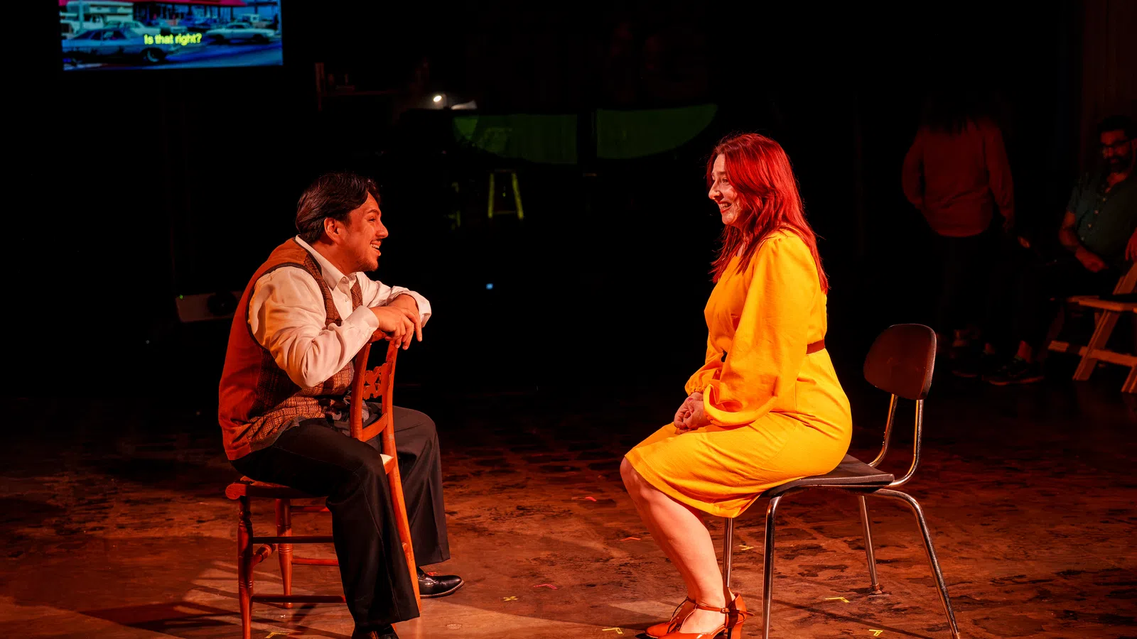 Man and woman in bright clothes sit facing each other on chairs in a dark theater setting with a screen in the background.