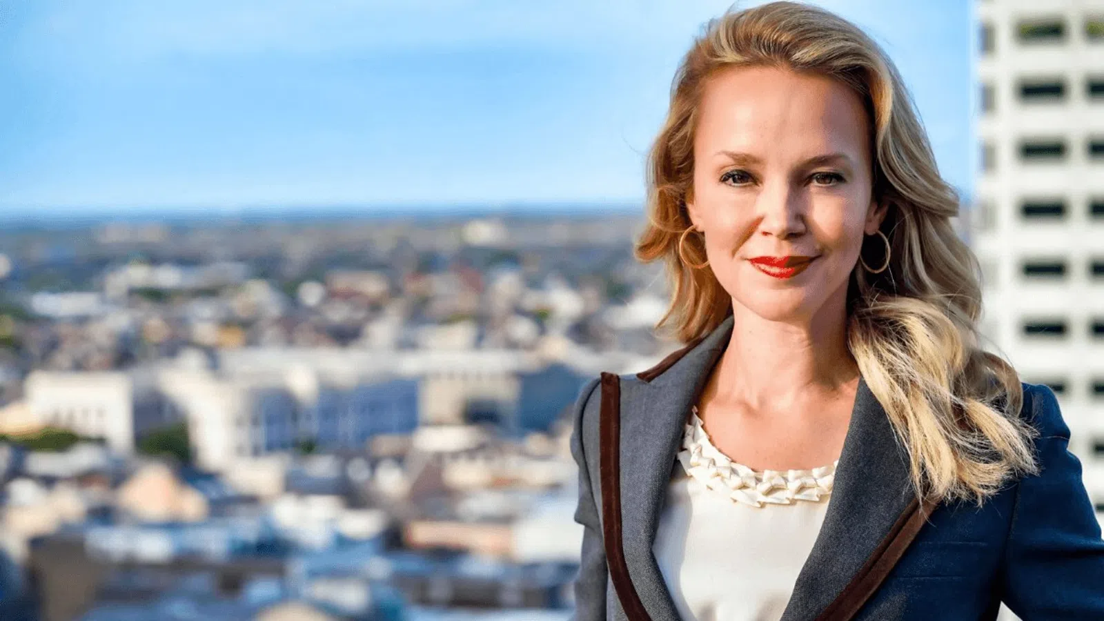 Professional woman with blonde hair smiling confidently in business attire against urban cityscape background.