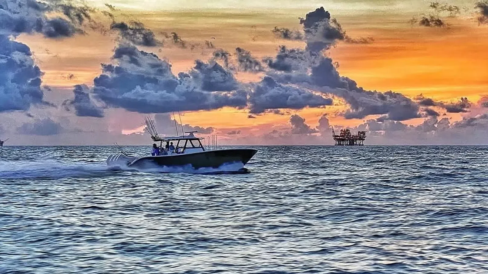 Speedboat cruising on ocean water against a dramatic sunset with scattered clouds and an offshore platform in view.