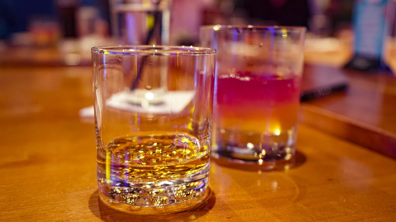 Two glasses on a wooden table, one with whiskey and the other with a layered cocktail, blurred background.