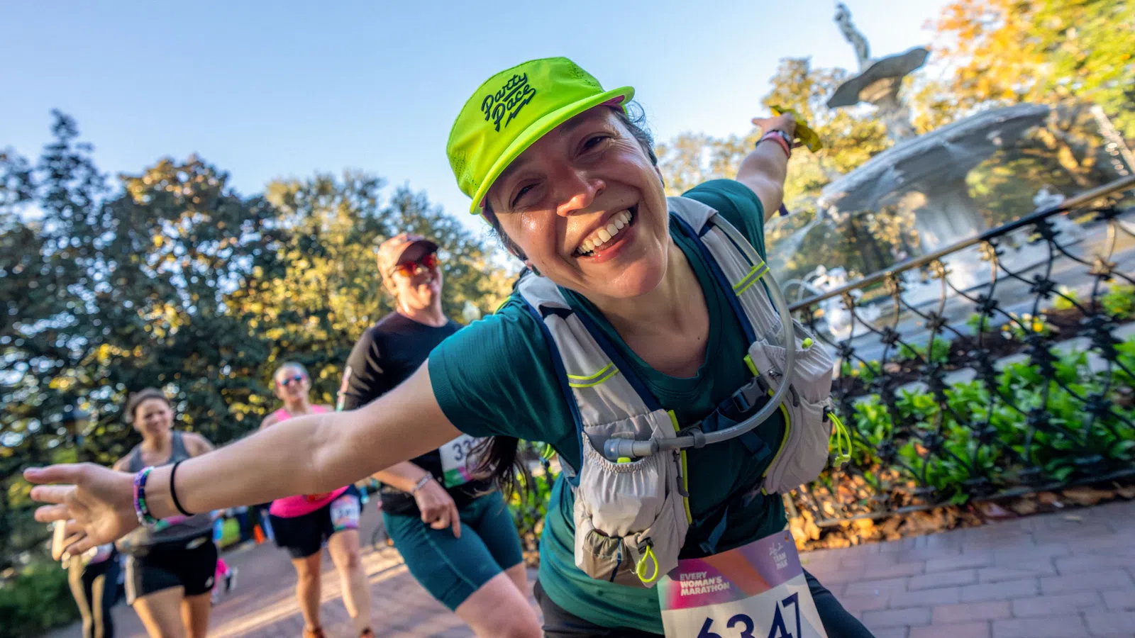 Smiling female marathon runner wearing neon green cap and bib 6347 gestures with arms wide on park path.