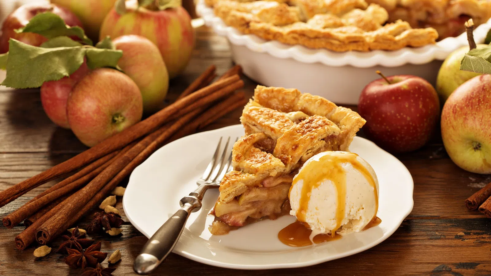 Slice of homemade apple pie with vanilla ice cream and caramel sauce on white plate with fresh apples and cinnamon sticks.