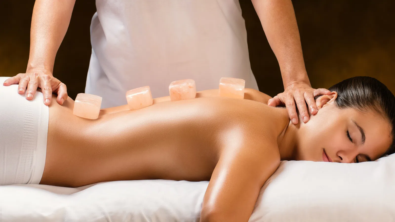 Woman receiving hot stone massage therapy with pink salt stones on her back in a spa setting.