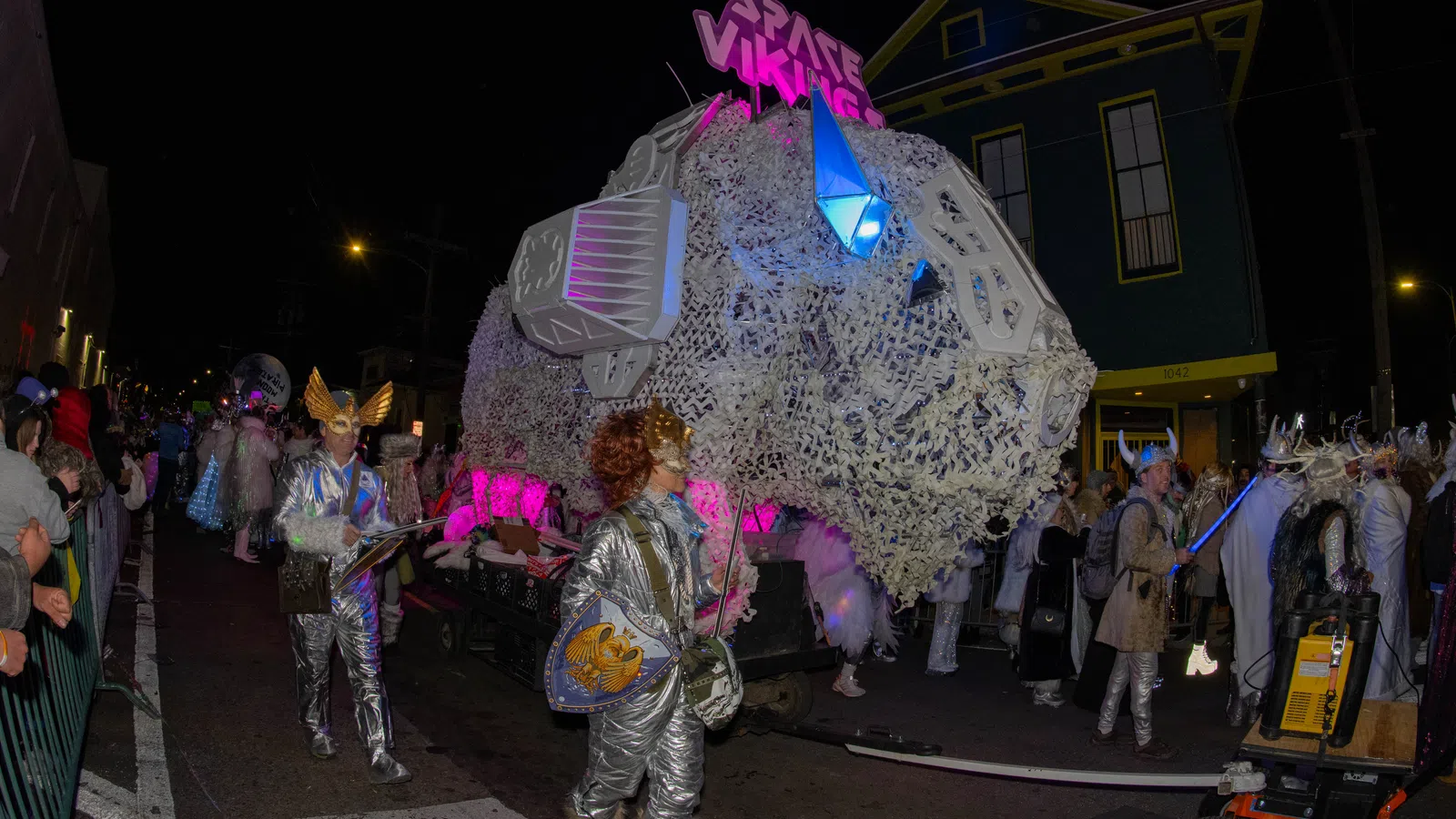 People in silver space-themed costumes parade beside a large illuminated float reading Space Vikings at night.