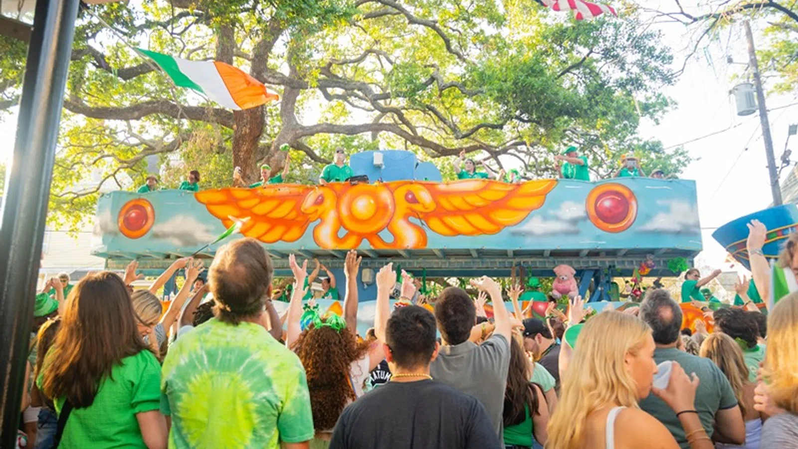 Crowd wearing green celebrates next to colorful parade float with Irish and American flags during festive event.