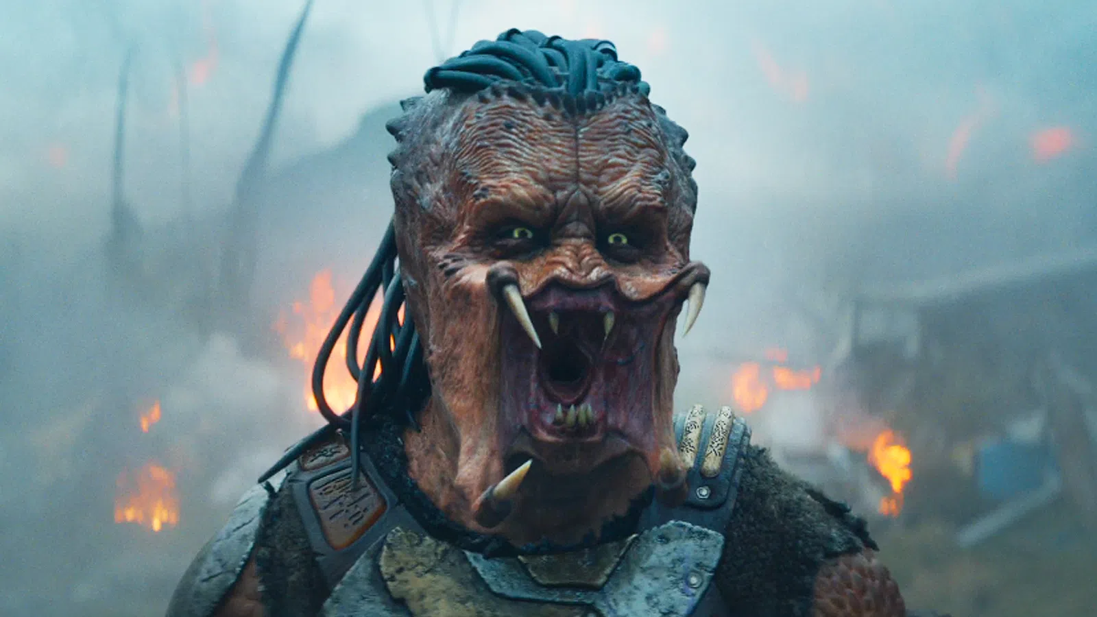 Close-up of a fierce alien creature with sharp teeth, dreadlock-like hair, and armor in smoky battlefield.
