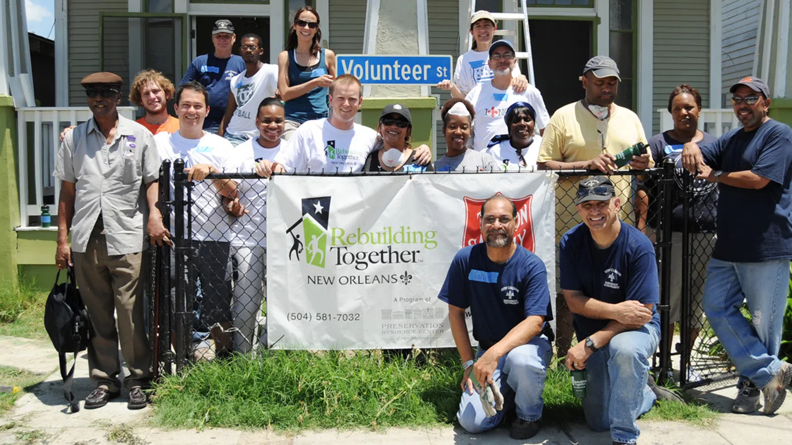 Group of diverse volunteers posing behind a Rebuilding Together New Orleans banner on a sunny day