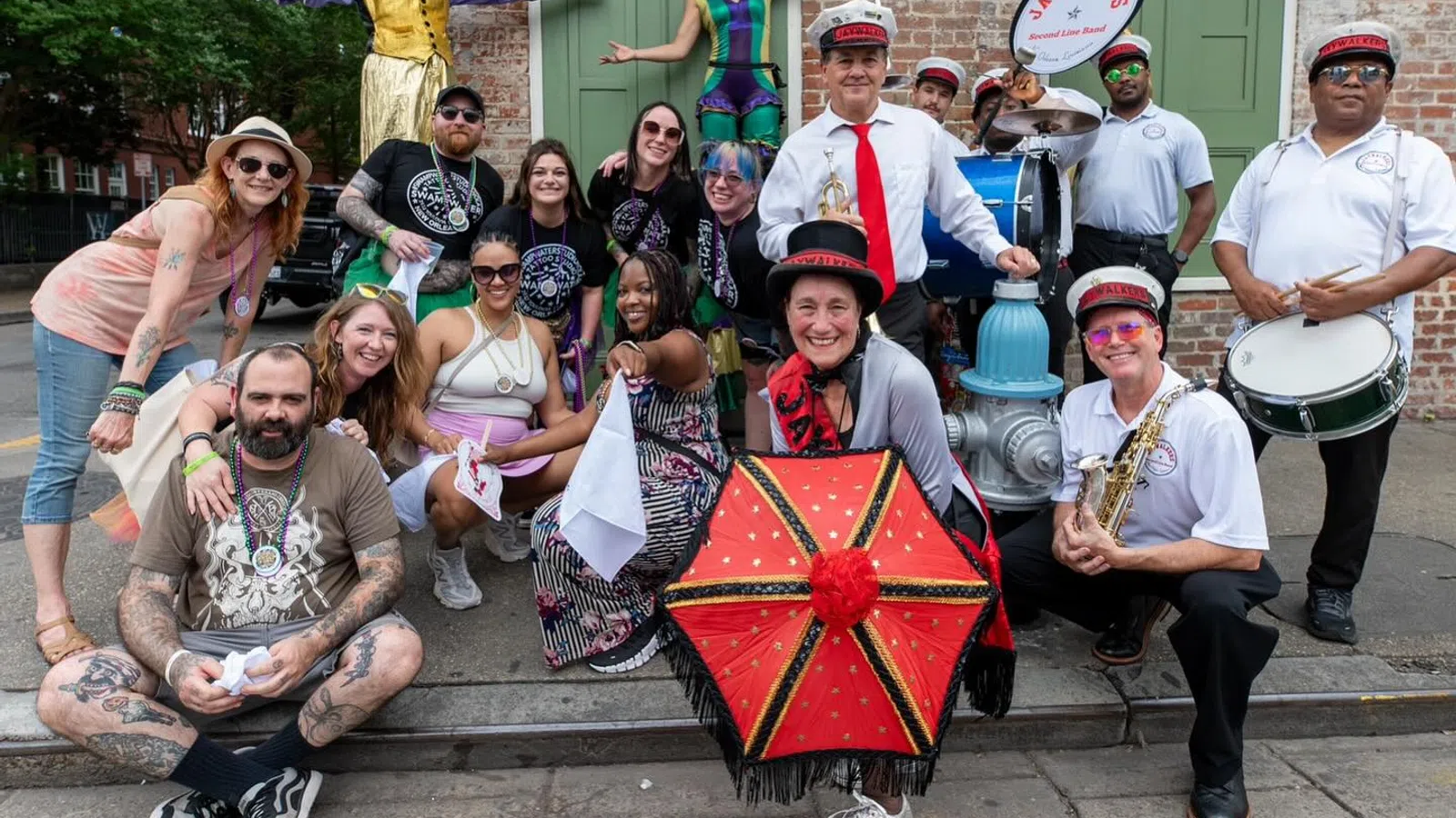 Group of diverse people and musicians posing joyfully outdoors with colorful costumes, instruments, and a decorated umbrella.