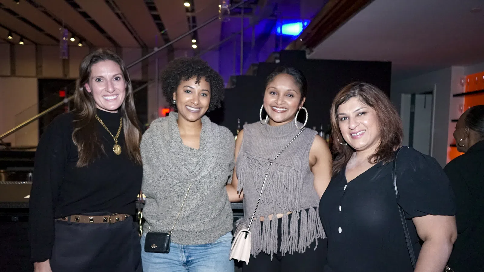Four diverse women smiling and posing together at an indoor event with modern lighting and decor.