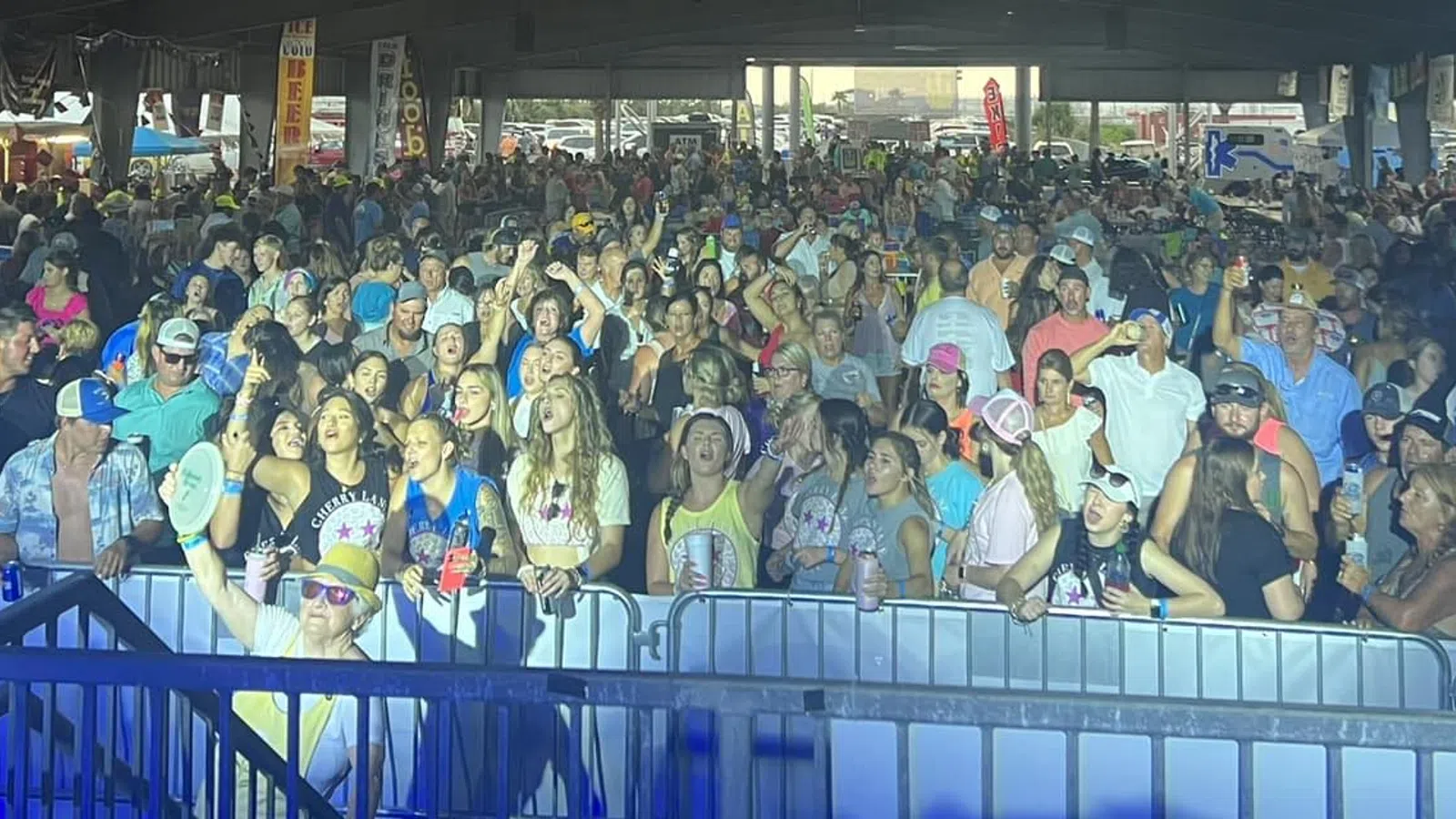 Large crowd enjoying an indoor concert or event with people cheering and taking photos under overhead lighting.