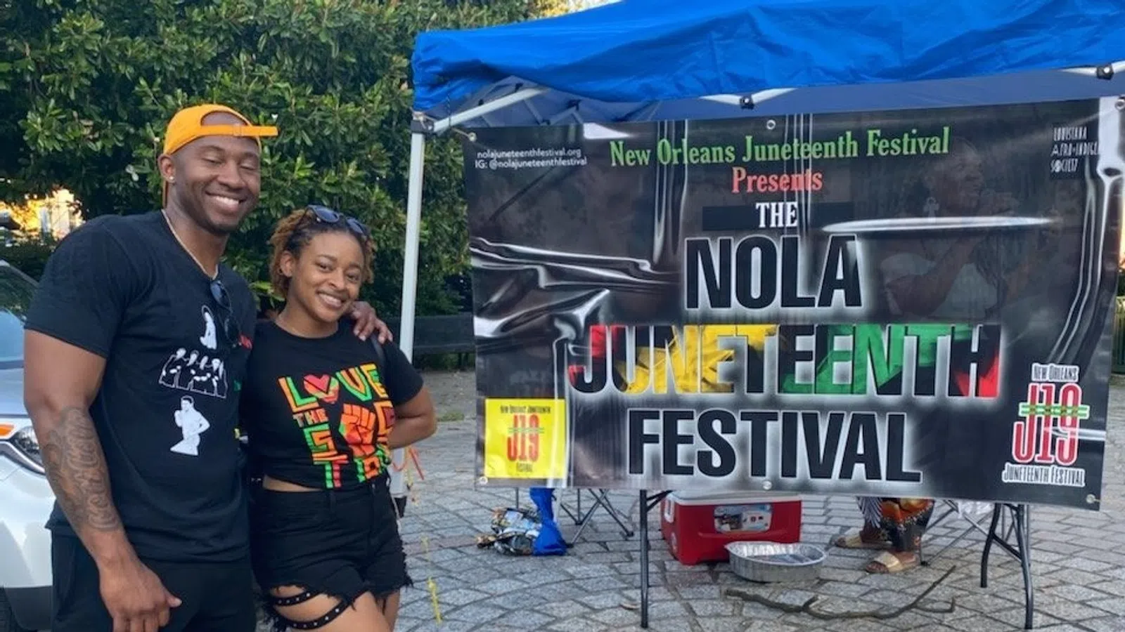 Smiling couple posing in front of a colorful NOLA Juneteenth Festival banner under a blue canopy on a cobblestone path.