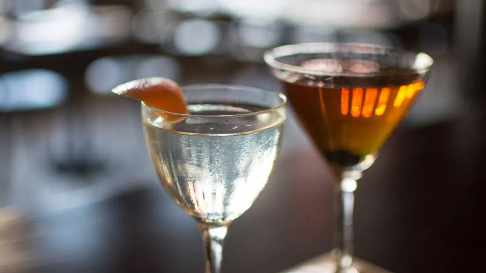 Two elegant cocktails on a bar counter, one clear with a citrus garnish and one amber-colored.
