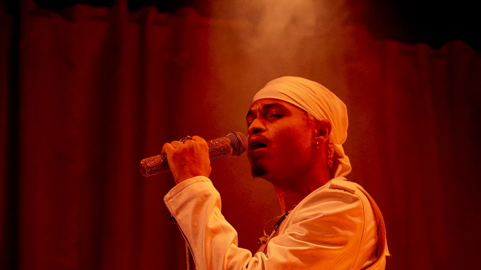 Male singer wearing a white headscarf and jacket performing with a microphone on stage under warm red lighting.