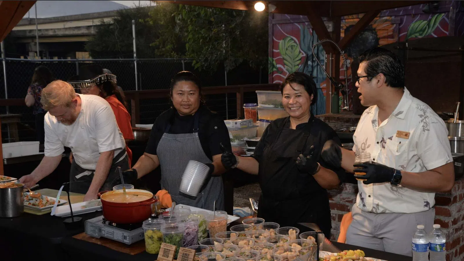 Four chefs preparing and serving a variety of dishes at an outdoor food stall under a wooden roof.