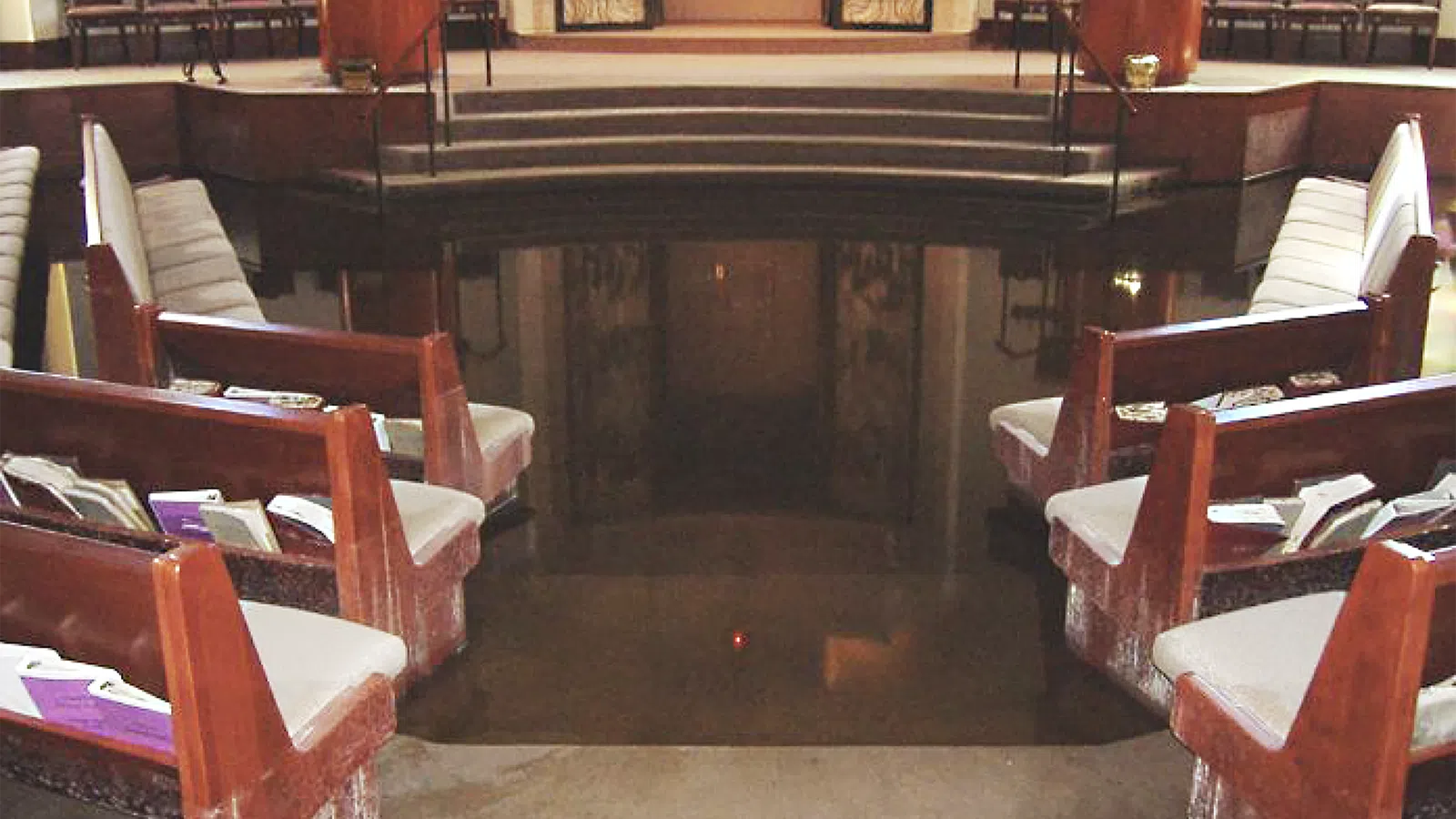 Interior of a synagogue with wooden benches, chairs, and a flooded floor reflecting the surroundings.