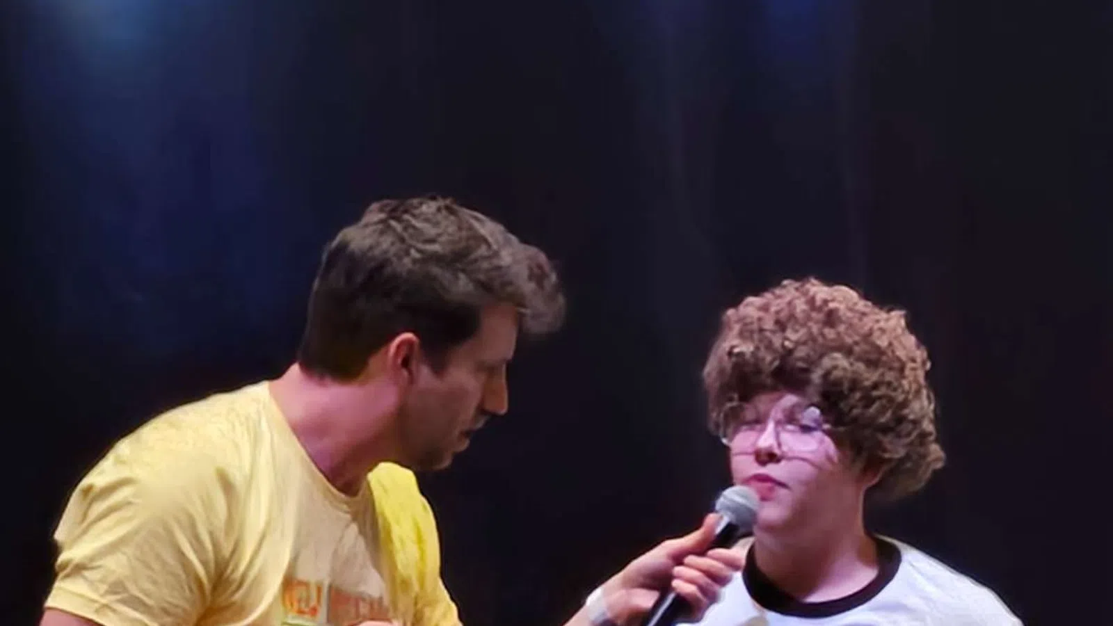 Two men on stage during a performance, one interviewing the other wearing a Vote for Pedro shirt and curly wig.