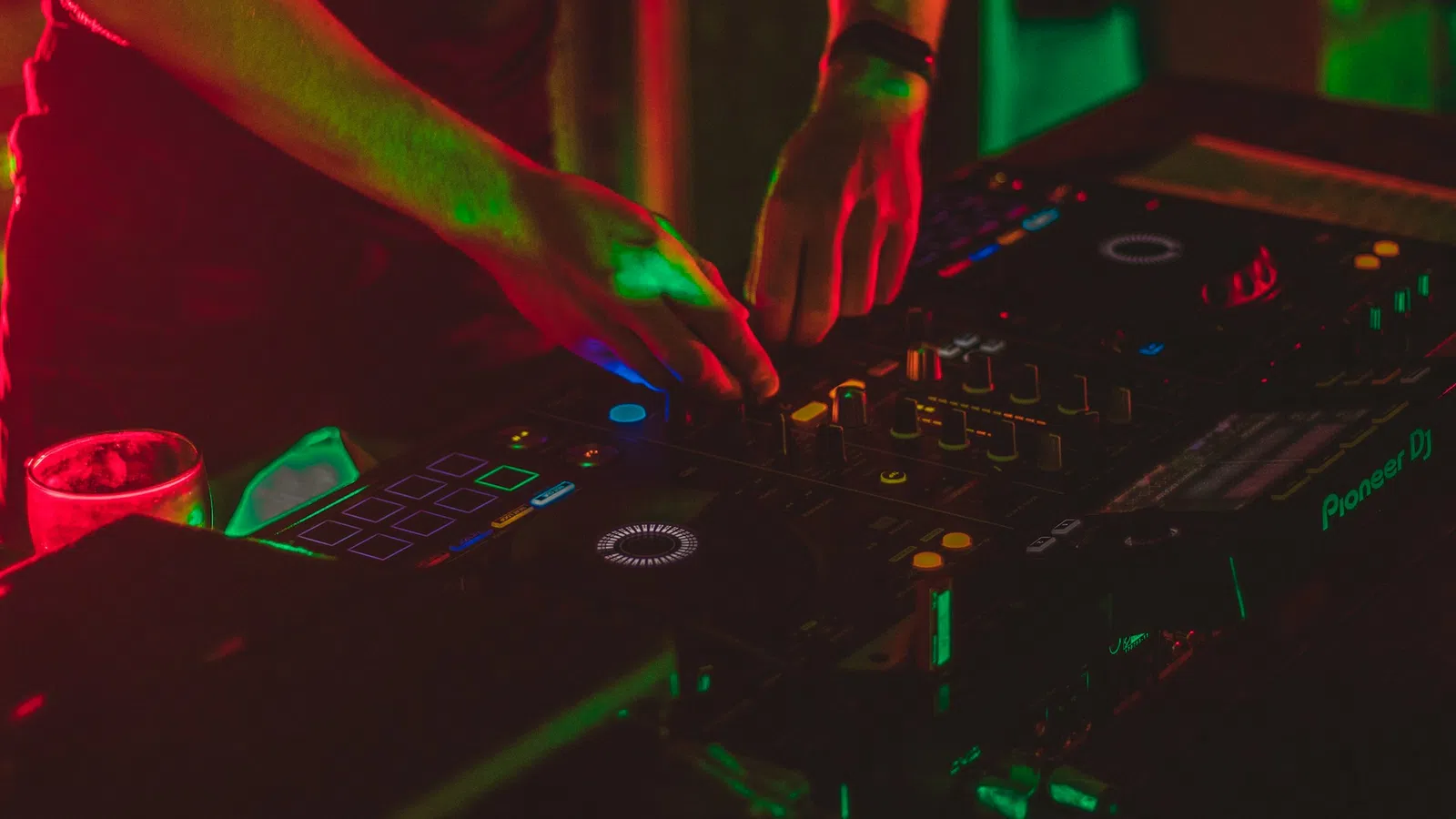 DJ mixing music on a Pioneer DJ controller under vibrant red and green club lights at night.