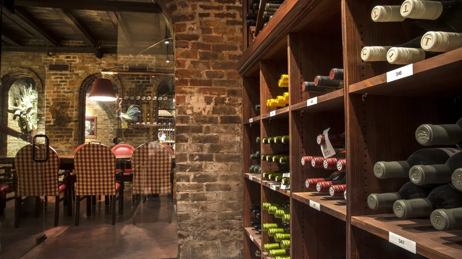 Wine bottles on wooden shelves in a dimly lit cellar dining room with exposed brick walls and checkered chairs.