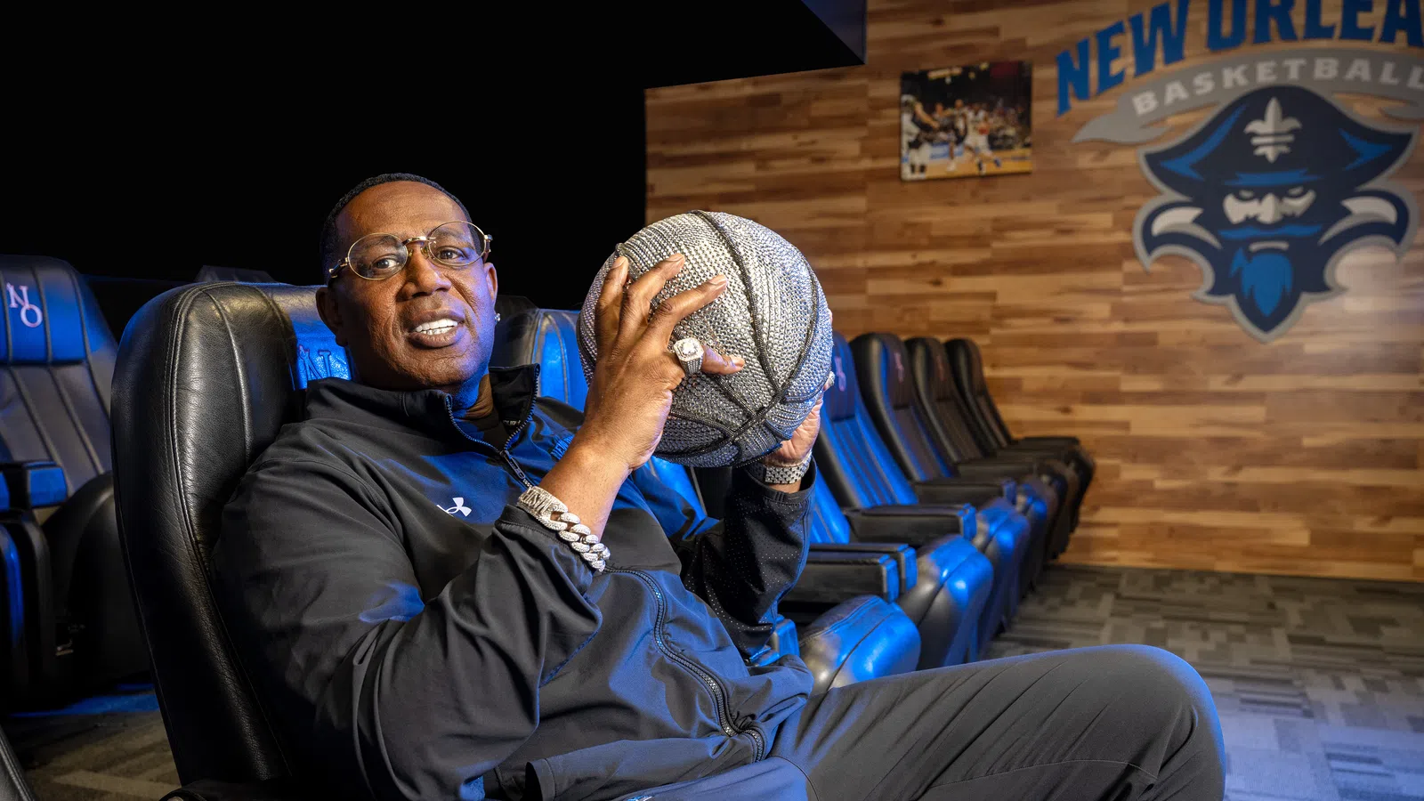 Man in black tracksuit holding a basketball sitting in a New Orleans Basketball lounge area with logo on wall.