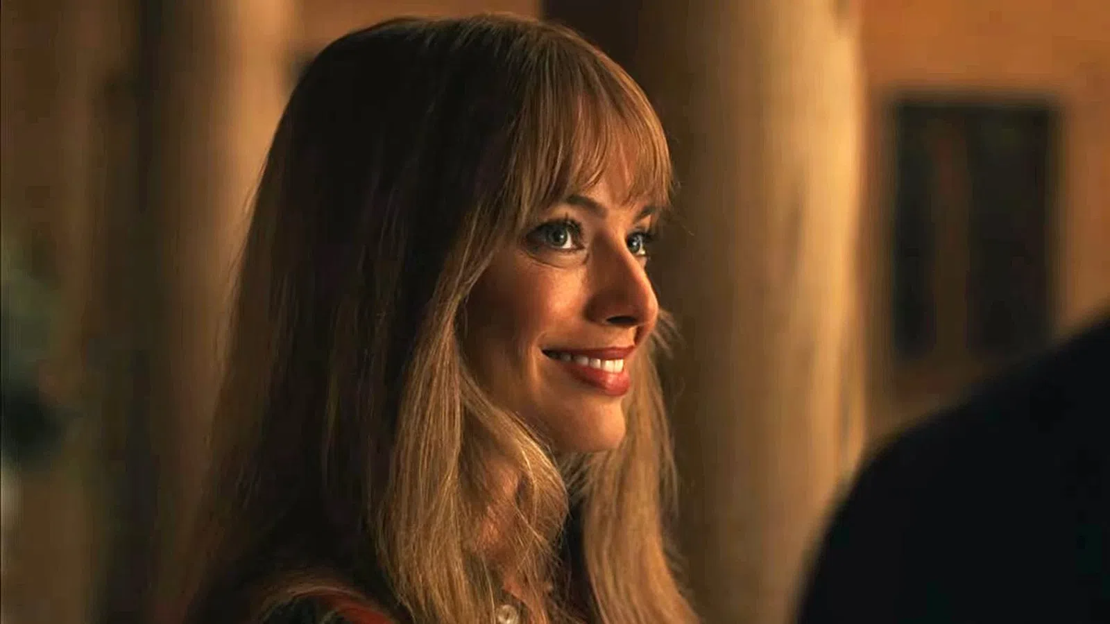 Smiling woman with long blonde hair and bangs looking to the side in warm indoor lighting.