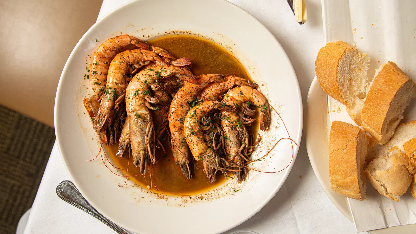 Plate of cooked shrimp in seasoned sauce served with slices of crusty bread on a white tablecloth.