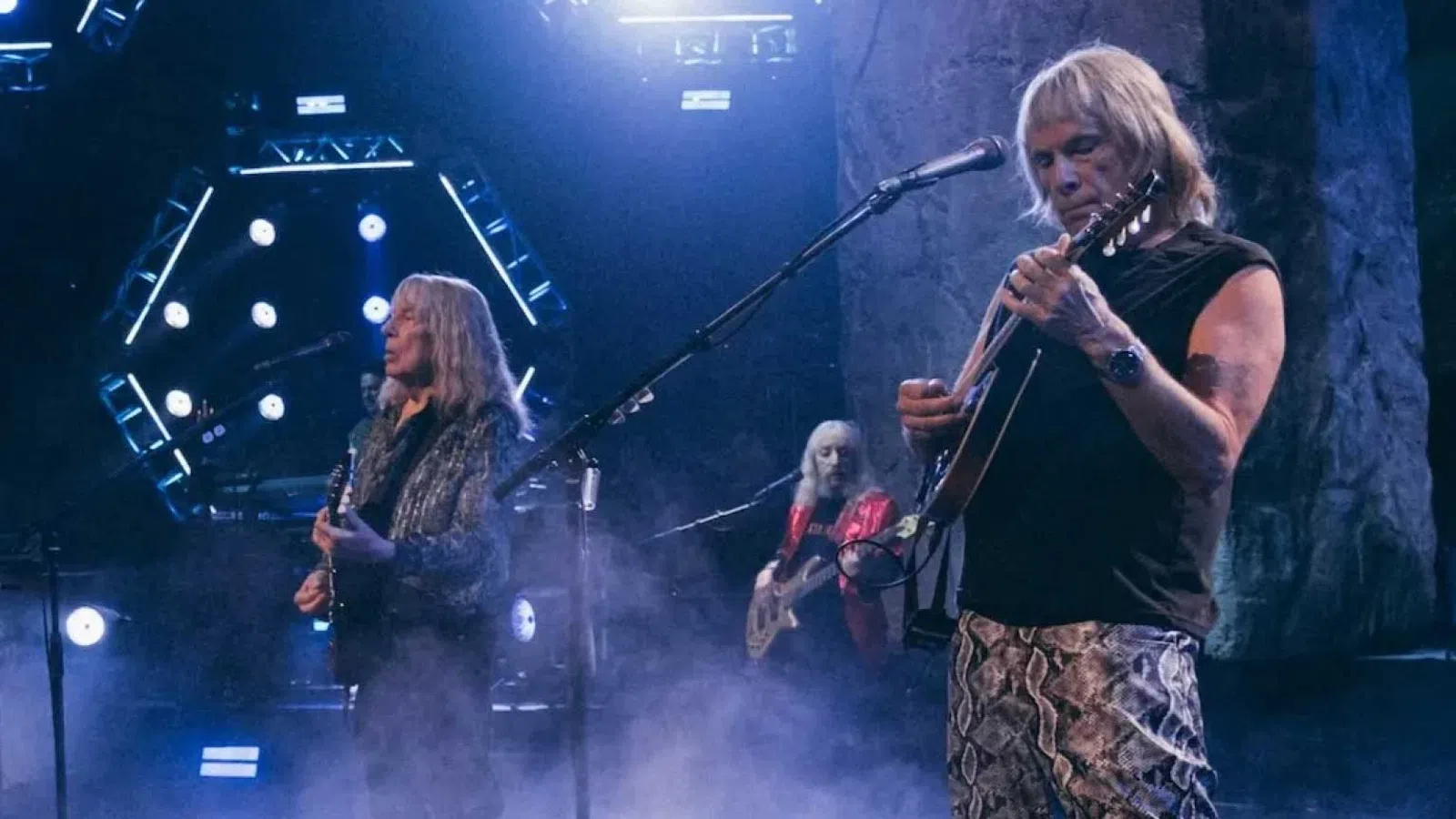 Rock band performing live on stage with guitars, piano, and dramatic lighting effects and smoke.