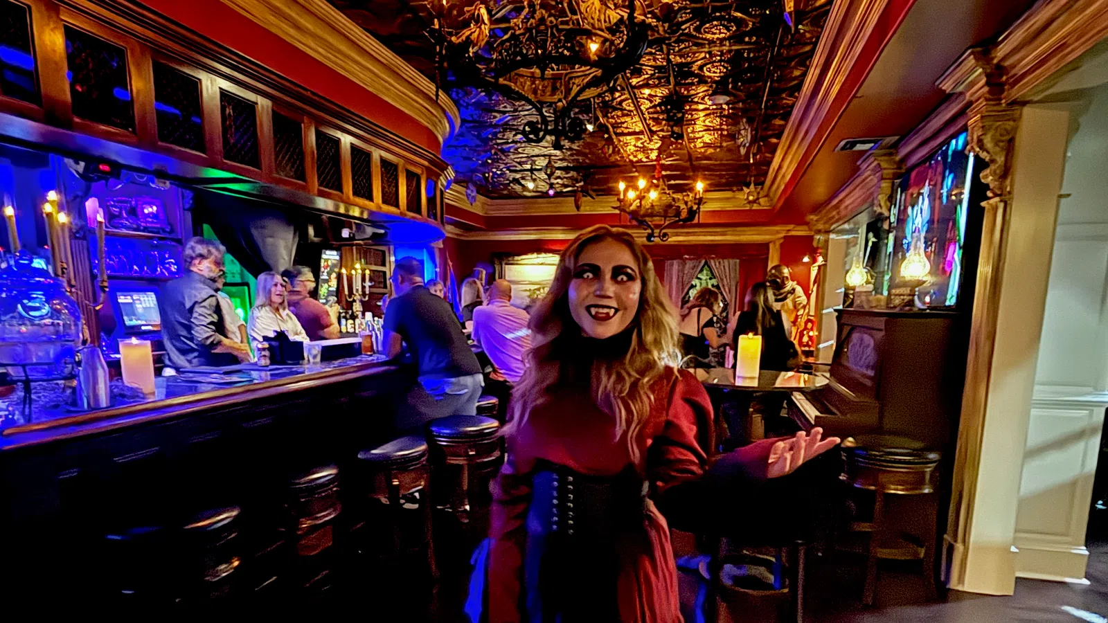 Woman in a red gothic dress with vampire makeup in a dimly lit ornate bar with patrons and warm chandeliers.