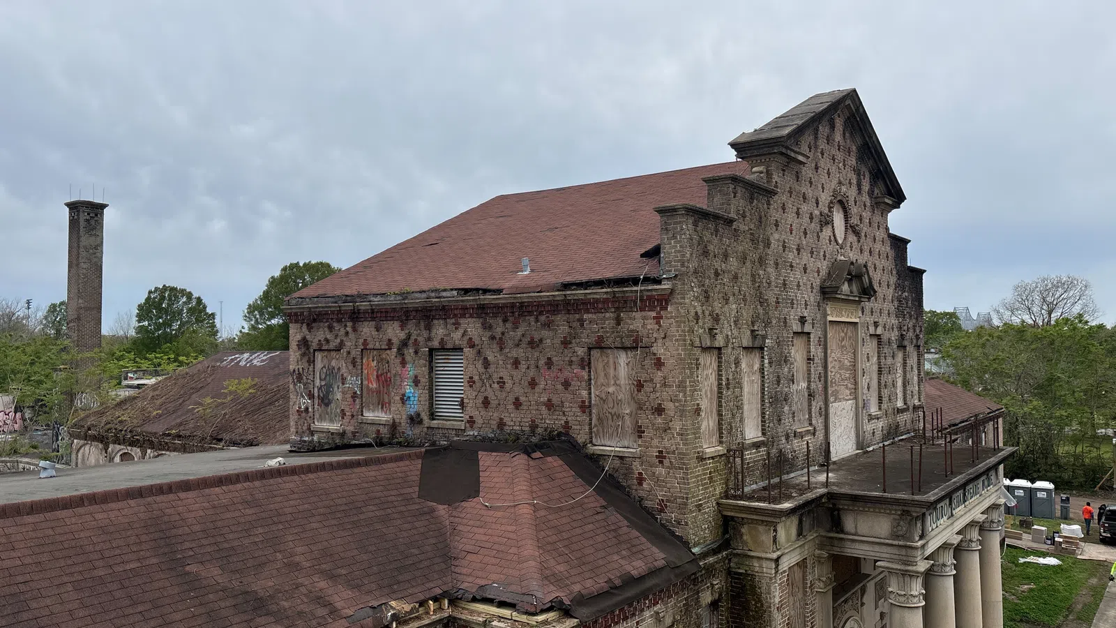 Abandoned brick building with boarded windows, red roof, and classical columns under a cloudy sky.