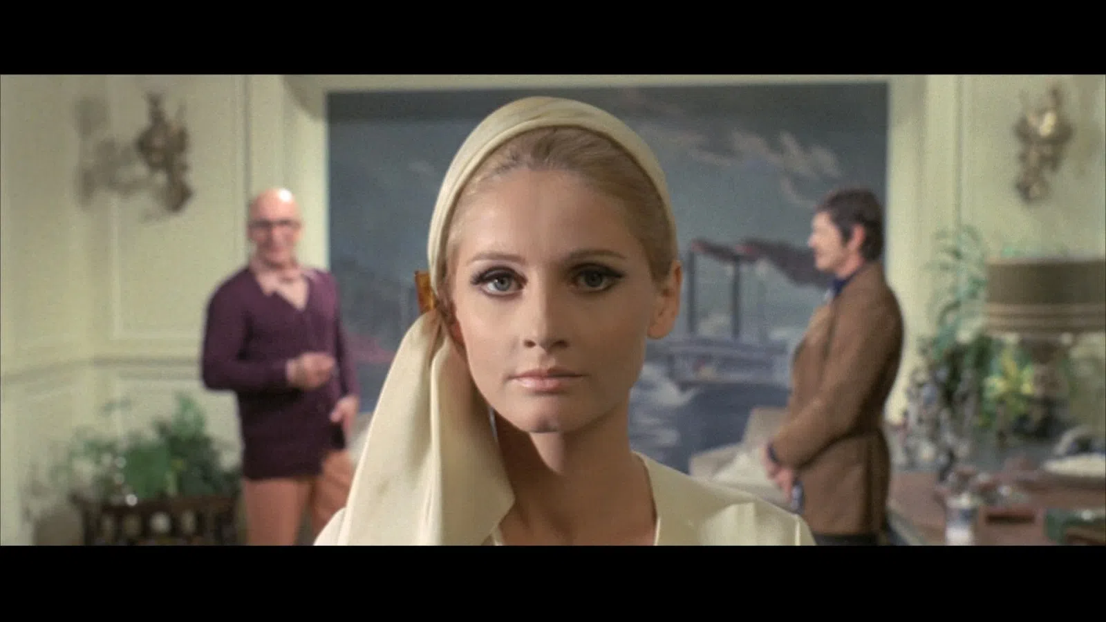 Close-up of a woman with makeup and headscarf, two men blurred in background inside a decorated room.