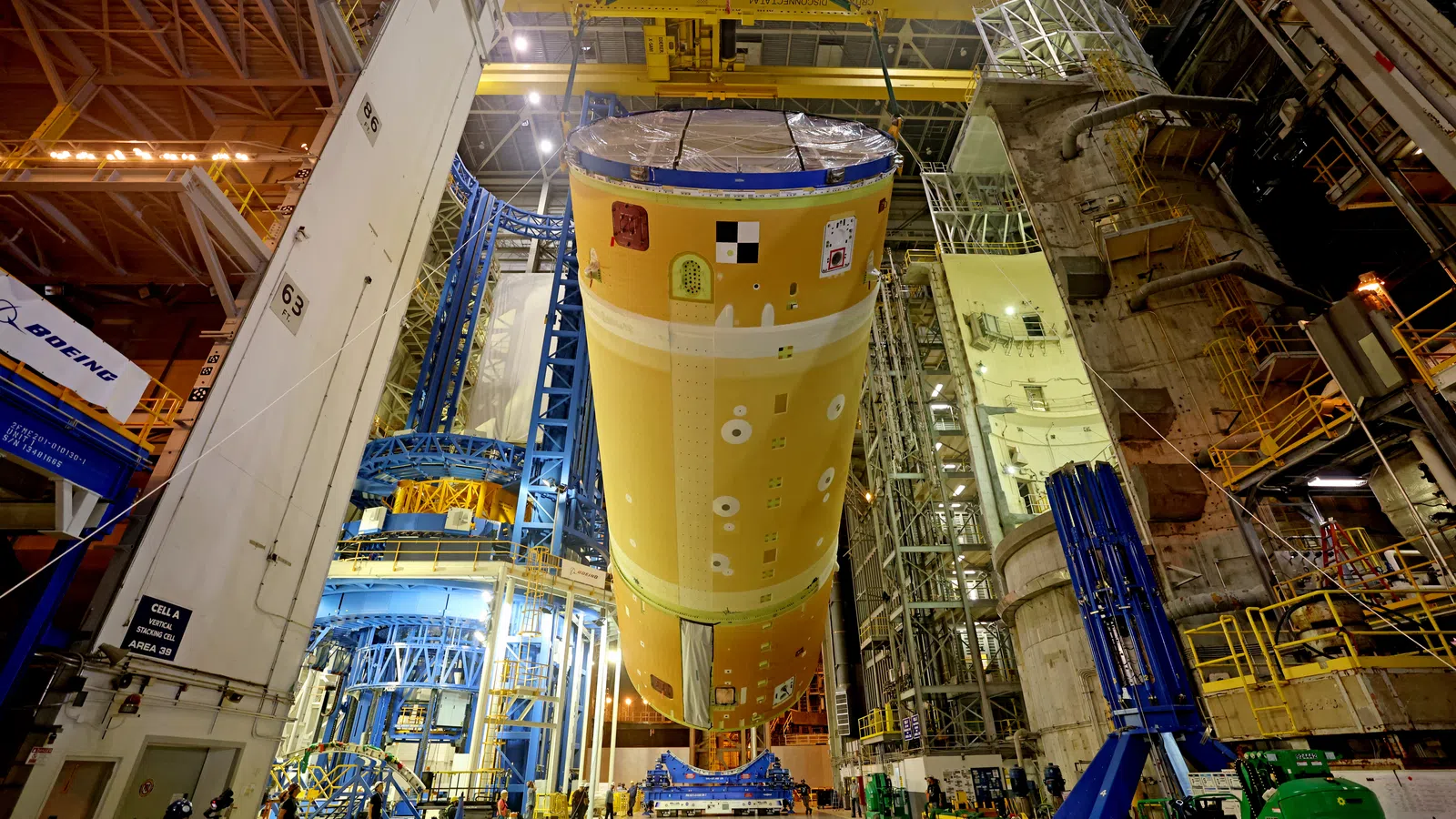 Large orange spacecraft fuel tank suspended inside a spacious industrial assembly facility with workers and machinery.
