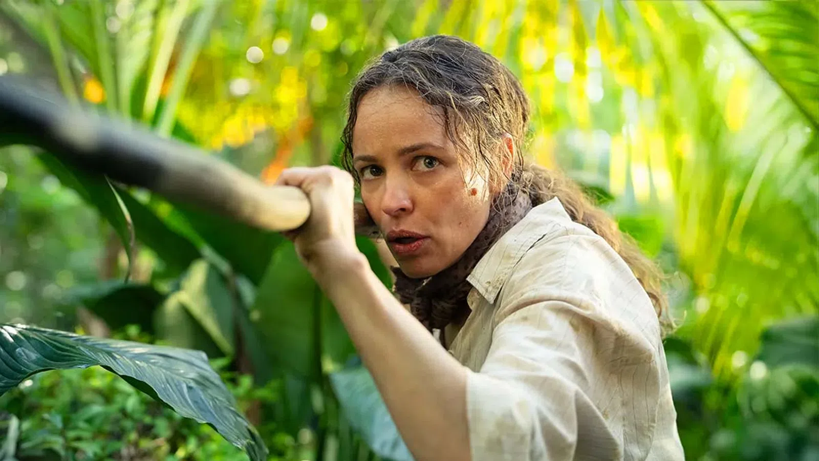 Woman in beige shirt cautiously holding a wooden stick amidst dense green jungle foliage.