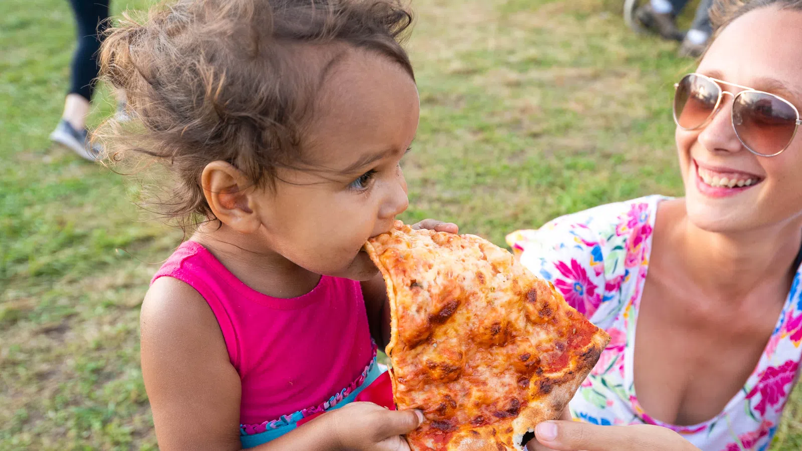 Young child wearing pink dress eating a large slice of pizza outdoors held by a smiling woman in sunglasses.