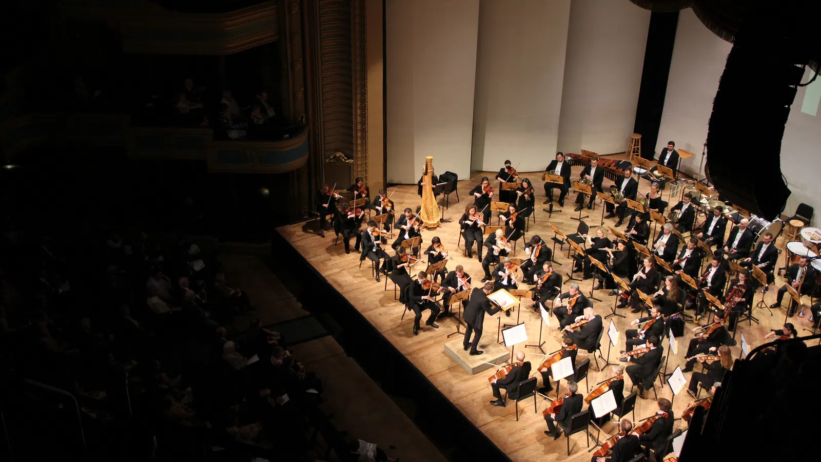 Full orchestra performing on stage in a concert hall with conductor leading musicians and audience seated in darkness