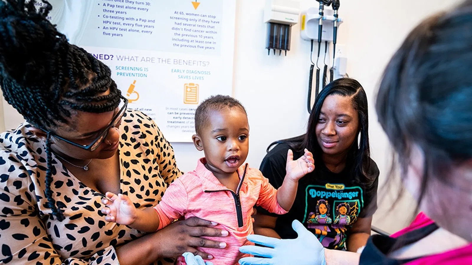 Child in pink shirt interacting with healthcare worker wearing gloves, accompanied by two women in a medical office.