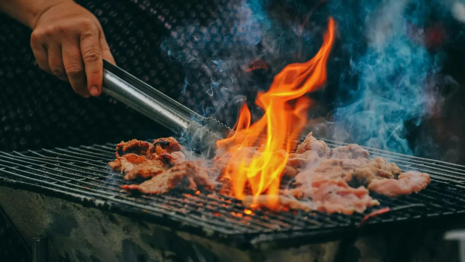 Hand grilling meat over an open flame with smoke rising from a charcoal barbeque grill outdoors.
