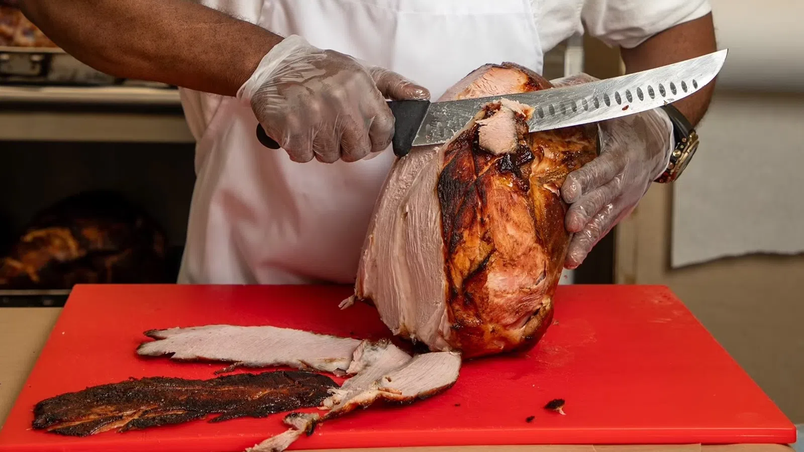 Chef slicing a large smoked ham on a red cutting board wearing gloves and white apron.