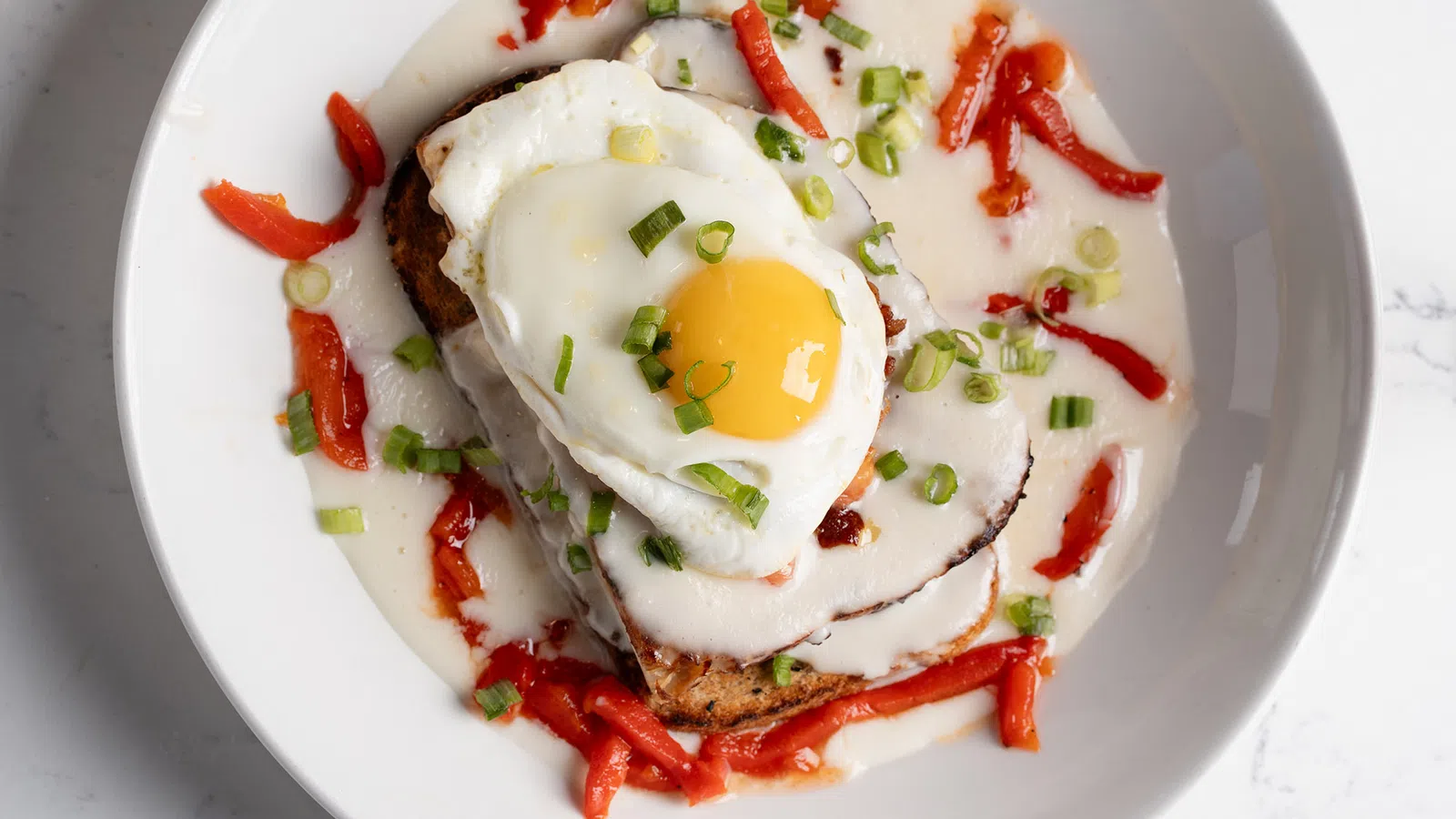 Fried egg atop toast with creamy sauce, red pepper strips, and chopped green onions on a white plate.