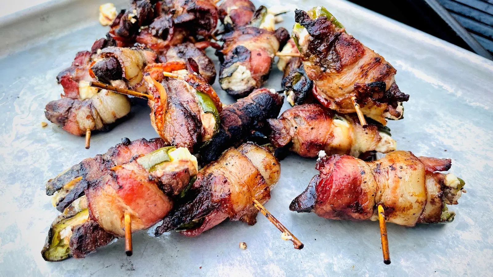 Grilled bacon-wrapped jalapeño poppers on a metal tray with toothpicks, showing charred and crispy textures.