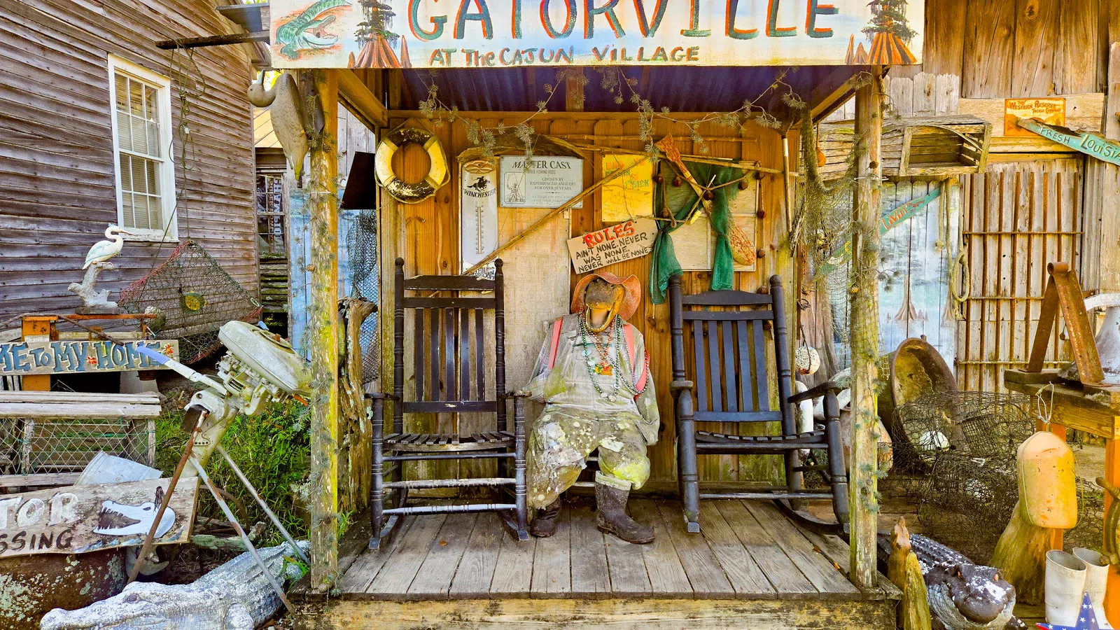 Rustic Gatorville porch with a scarecrow figure, rocking chairs, fishing gear, and vintage signs at Cajun Village.