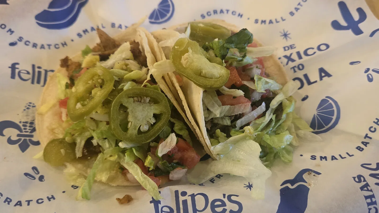 Two soft tacos filled with lettuce, jalapenos, tomatoes, and seasoned meat on branded paper wrapper.
