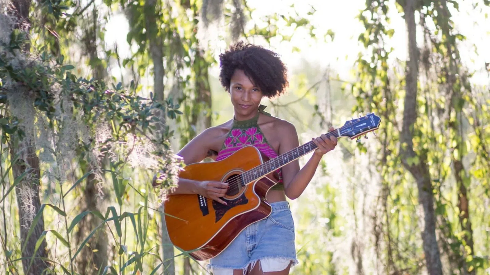 Young woman playing acoustic guitar barefoot in a sunny, lush forest clearing with hanging moss and sand.