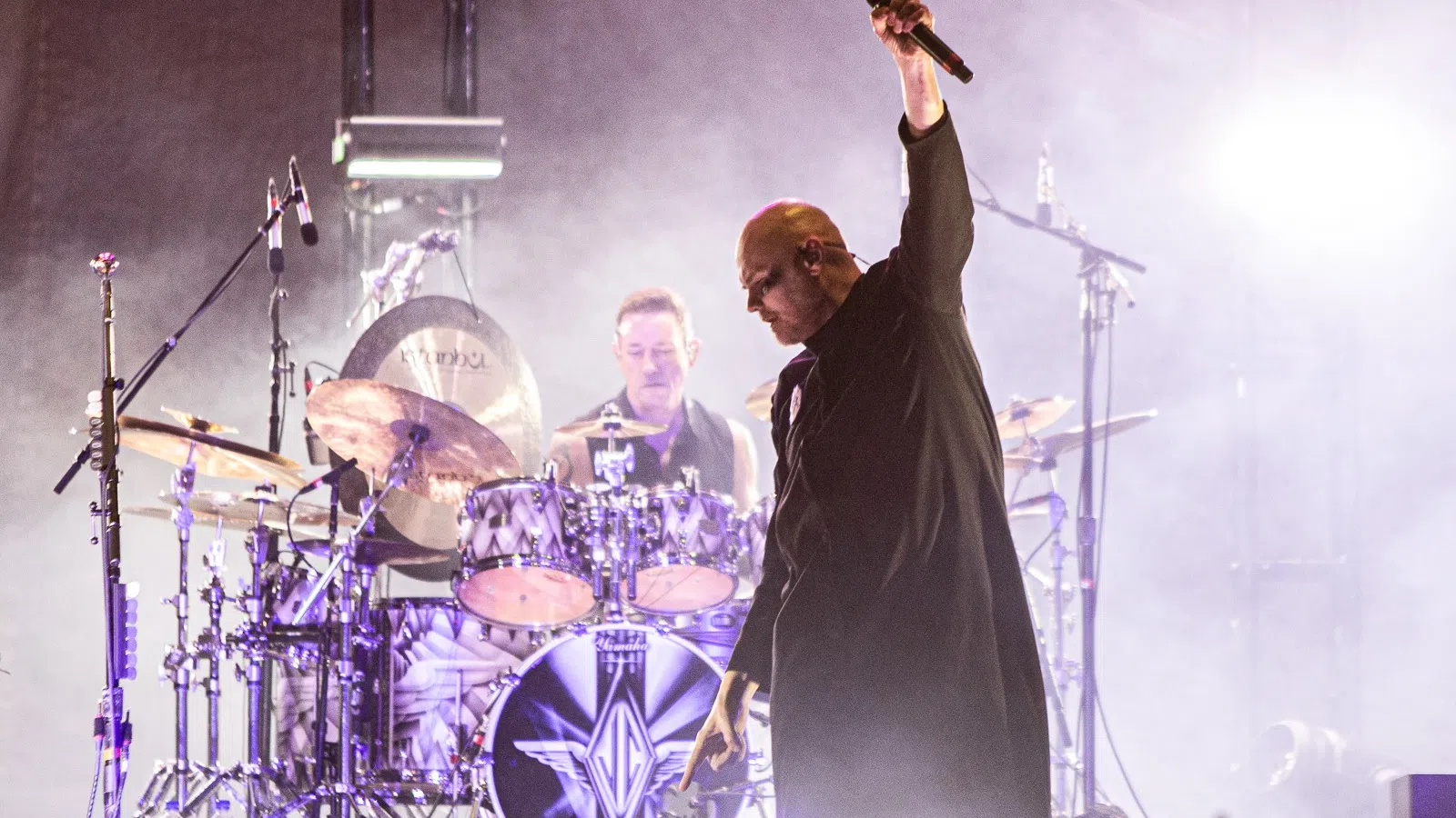 a person holding a microphone and standing next to a person playing drums