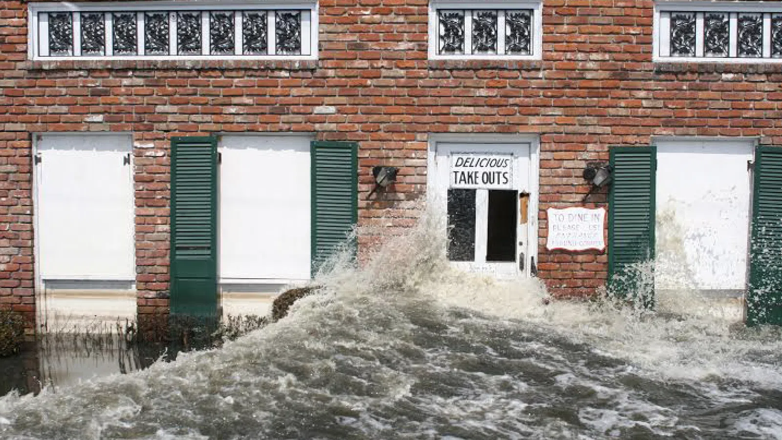 Floodwaters surge against a brick building facade with closed windows and a takeout restaurant door.