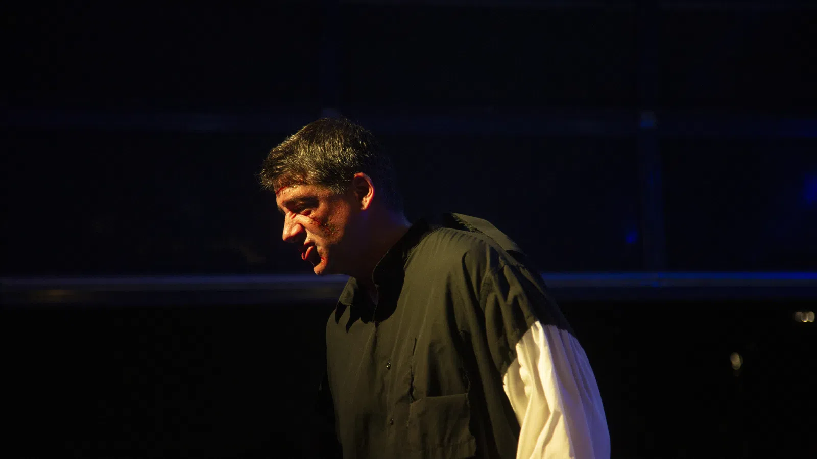 Man in dramatic lighting wearing black shirt with white sleeves, performing or speaking intensely on stage